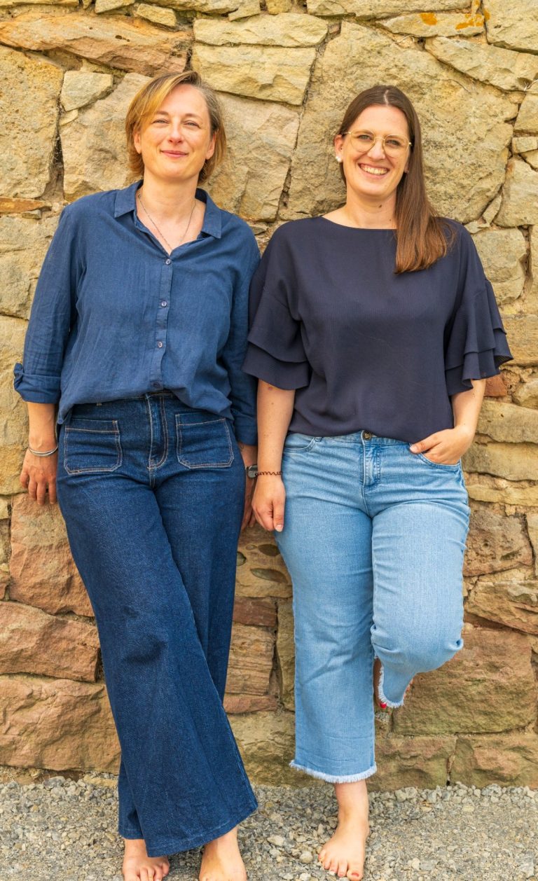 Zwei Frauen stehen lässig vor einer Steinmauer, beide in Jeans und Oberteilen.