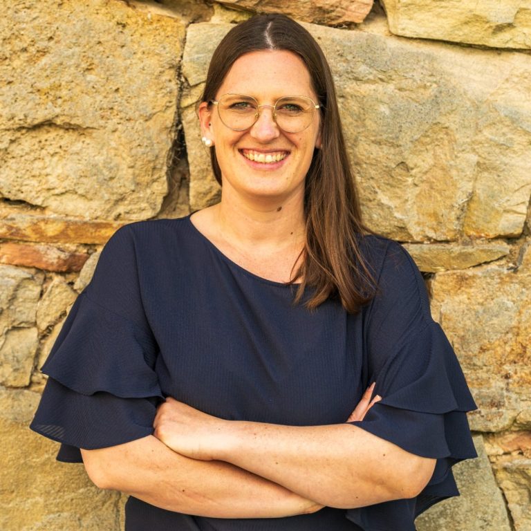 Lächelnde Frau mit langen Haaren, Arme vor der Brust verschränkt, vor einer steinernen Wand.
