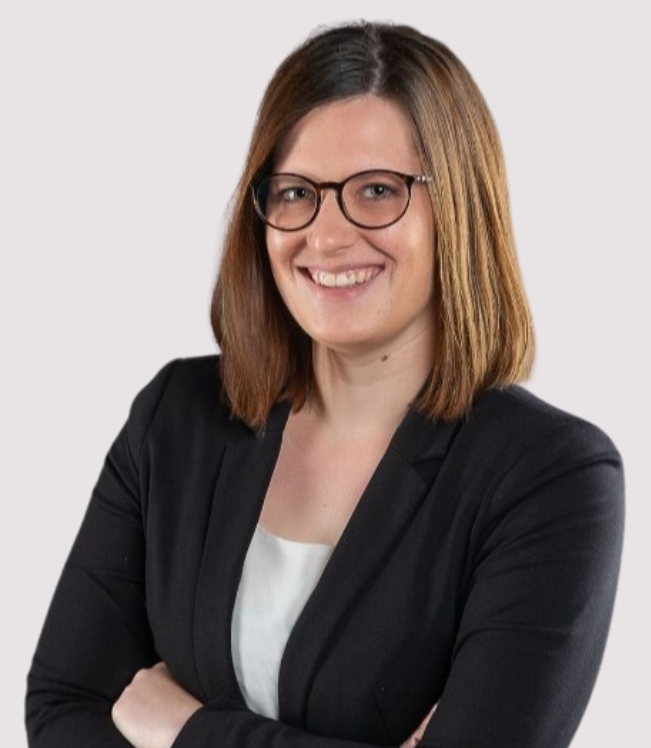 Kathrin Heimburger Junge Frau mit Brille, grauem Oberteil und Mappe, lächelt vor einer Wand.