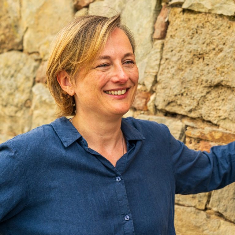 Lächelnde Frau in blauer Bluse vor einer Steinmauer.